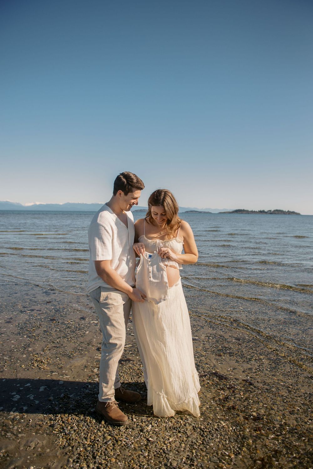Couple in white outfits standing on a rocky beach shore, holding baby clothes, with calm ocean and mountains behind.