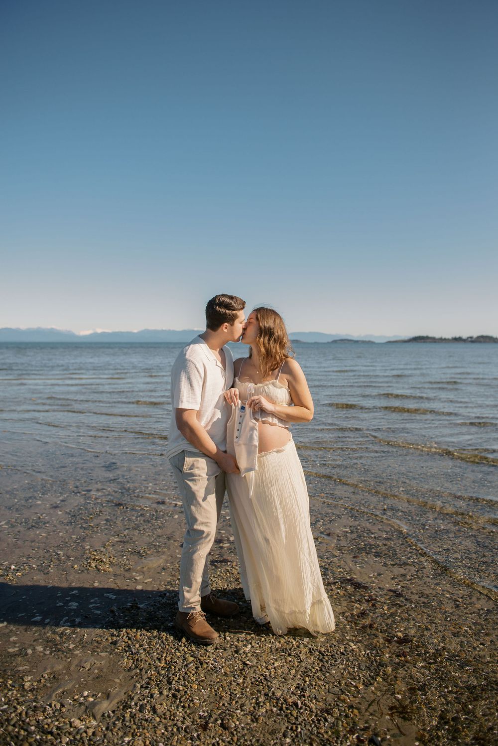 Couple sharing a kiss on a rocky beach shore with calm ocean waters and mountain views under a clear blue sky.