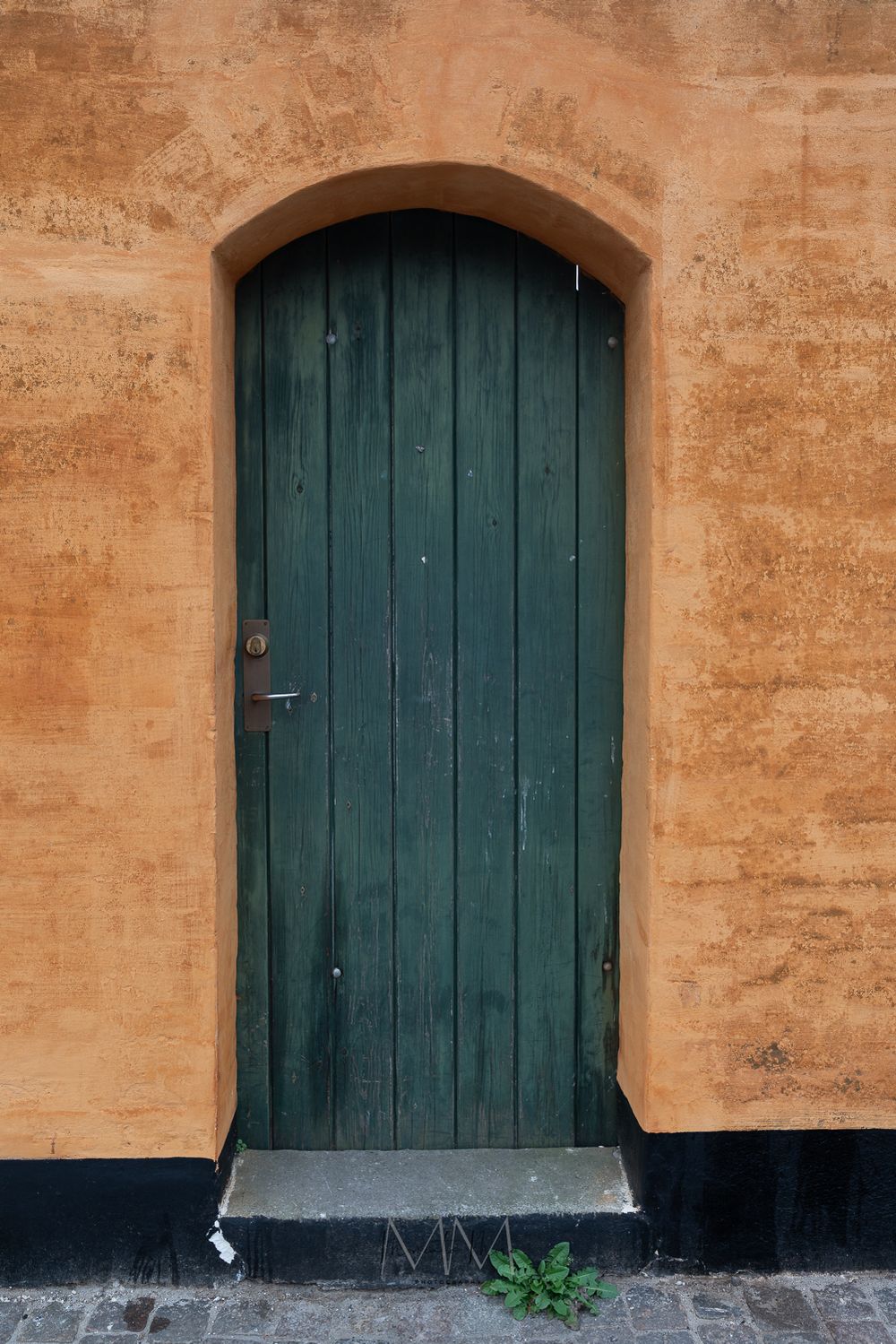 Rustic green arched wooden door set in an orange stucco wall with cobblestone steps.