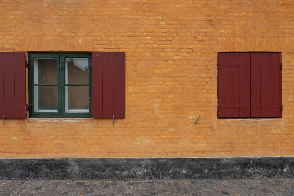 Orange brick building facade with two red wooden shuttered windows, one open and one closed.