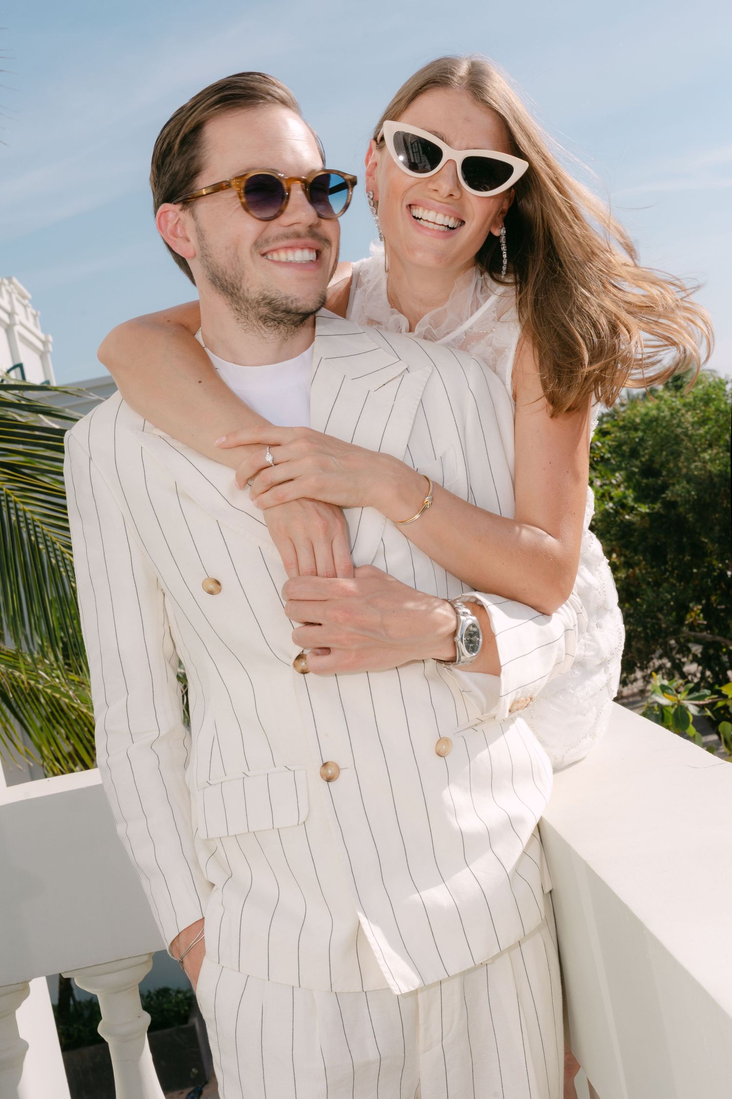 Couple in matching white pinstripe suits embracing on a sunny balcony, both wearing stylish sunglasses.