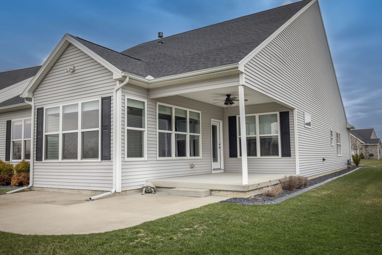 Single-story home with gray vinyl siding, covered patio, ceiling fan, black shutters, and well-maintained lawn.