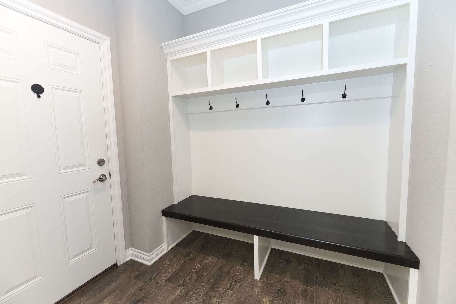 White built-in mudroom with open shelving, coat hooks, and a dark wood bench on hardwood floors near an entry door.