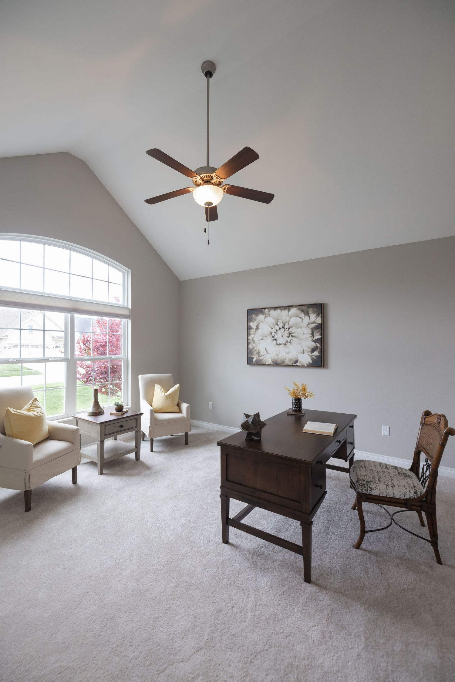 Bright home office with vaulted ceiling, ceiling fan, dark wood desk, cream armchairs, and large arched window with natural light.