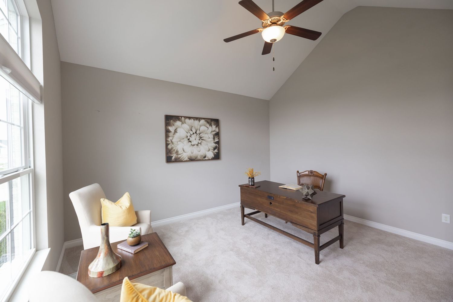 Bright living room with vaulted ceiling, ceiling fan, gray walls, carpet flooring, wooden console table, and cozy seating area.