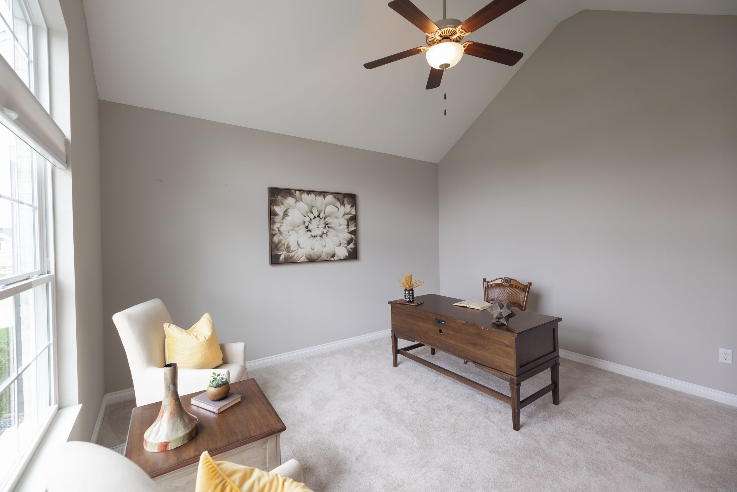 Bright living room with vaulted ceiling, ceiling fan, gray walls, carpet flooring, wooden console table, and cozy seating area.