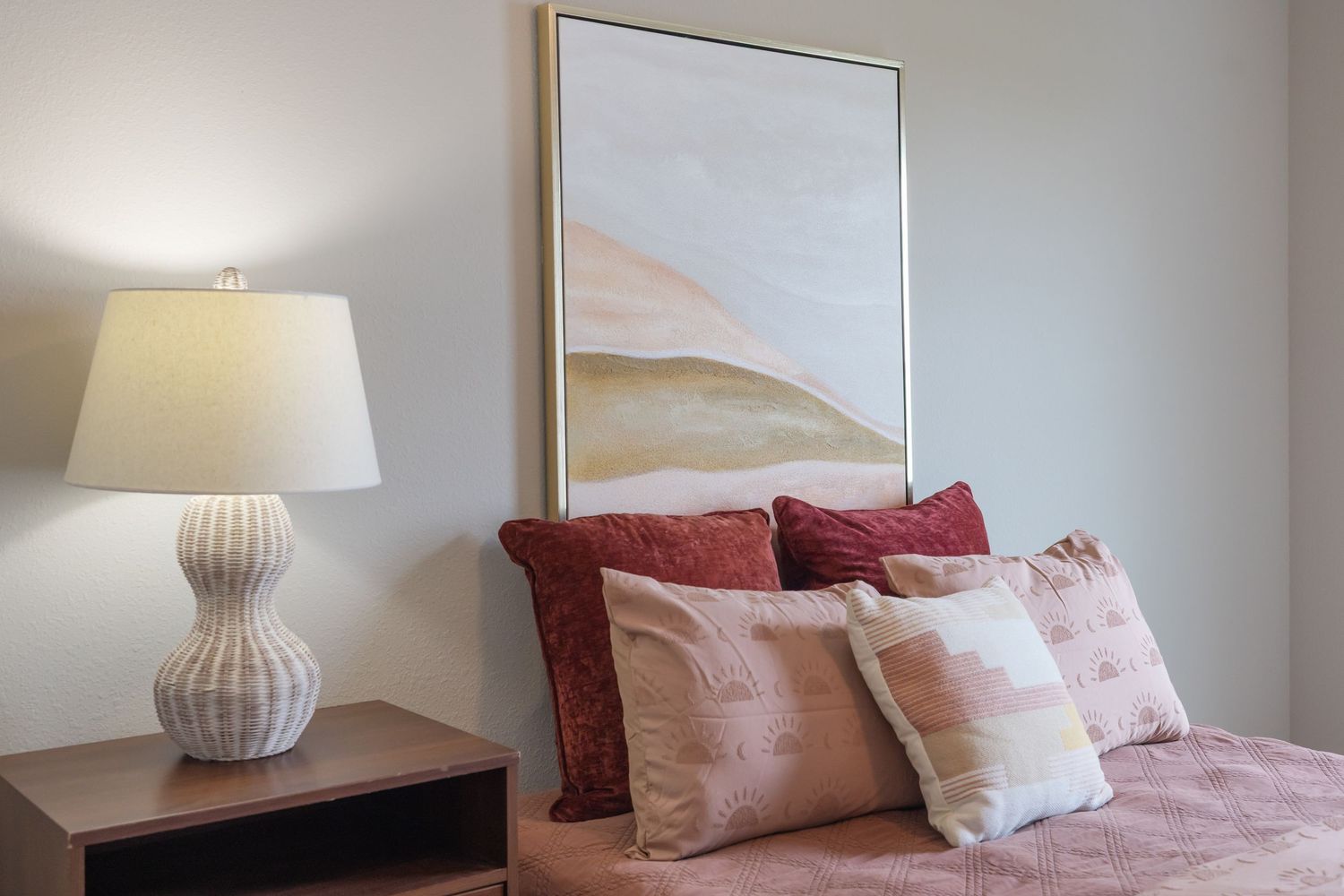 Cozy bedroom with pink bedding, decorative pillows, a table lamp, and abstract wall art.
