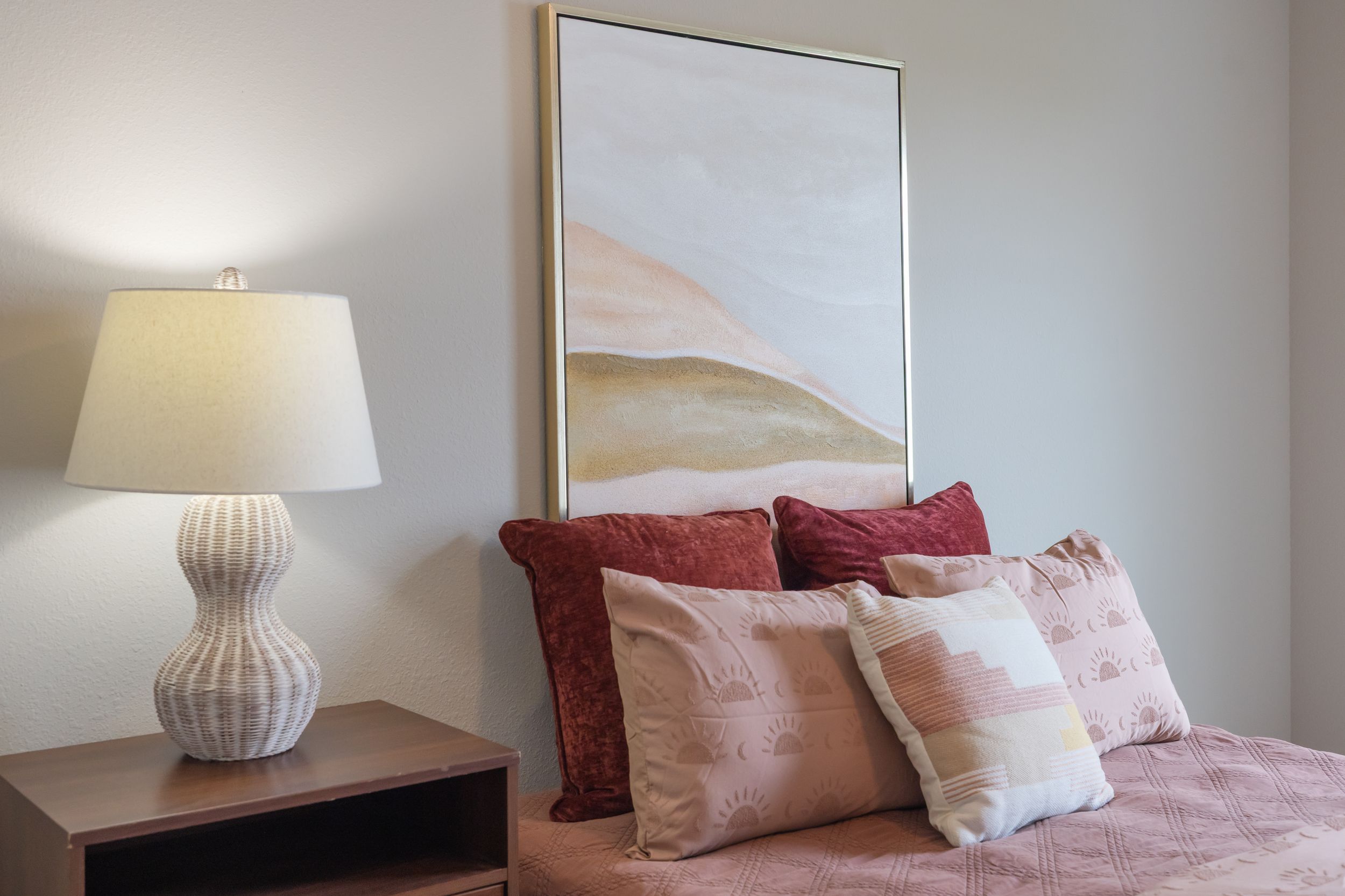 Cozy bedroom with pink bedding, decorative pillows, a table lamp, and abstract wall art.
