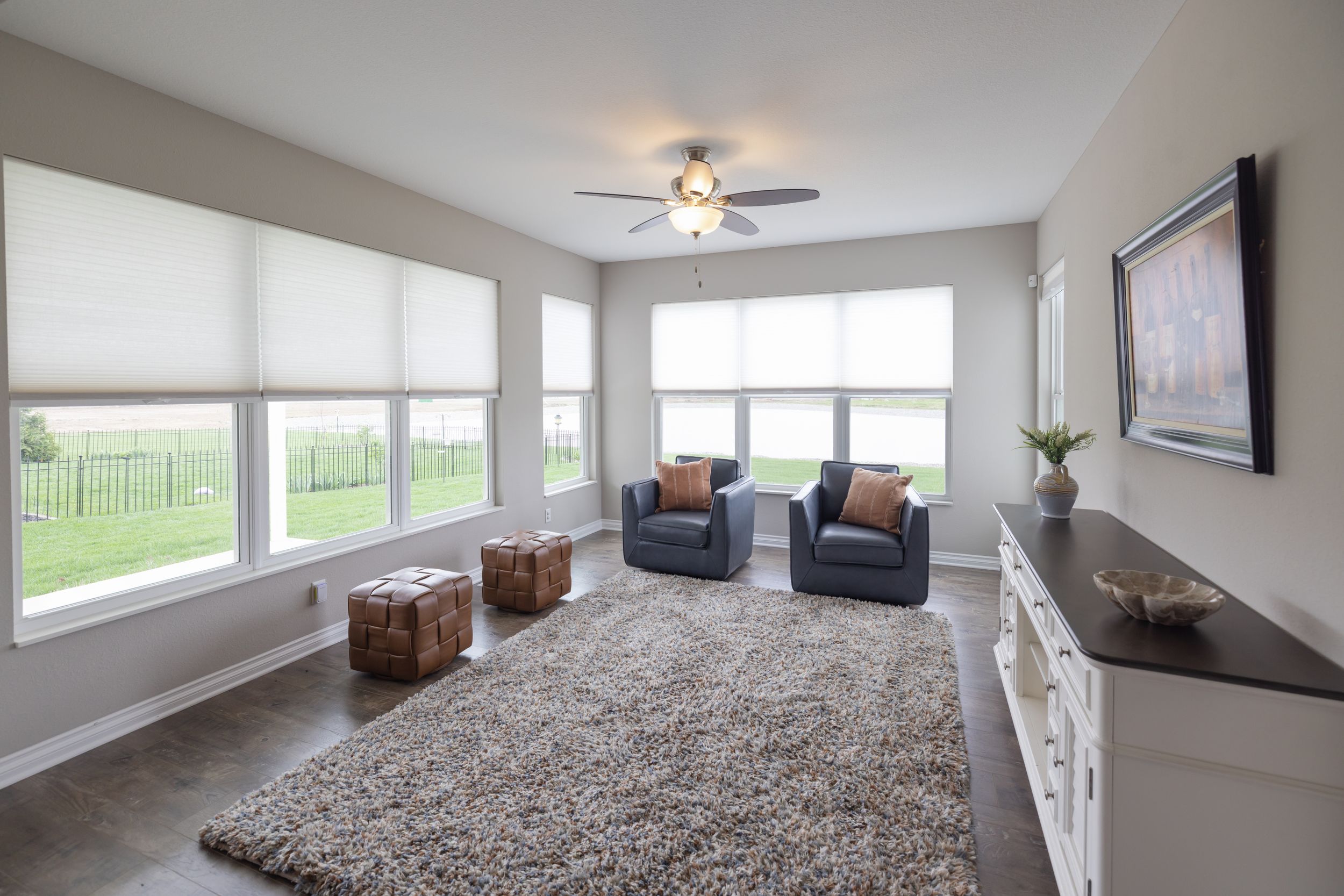 Bright living room with blue armchairs, shag rug, roller shades on large windows, ceiling fan, and white dresser.