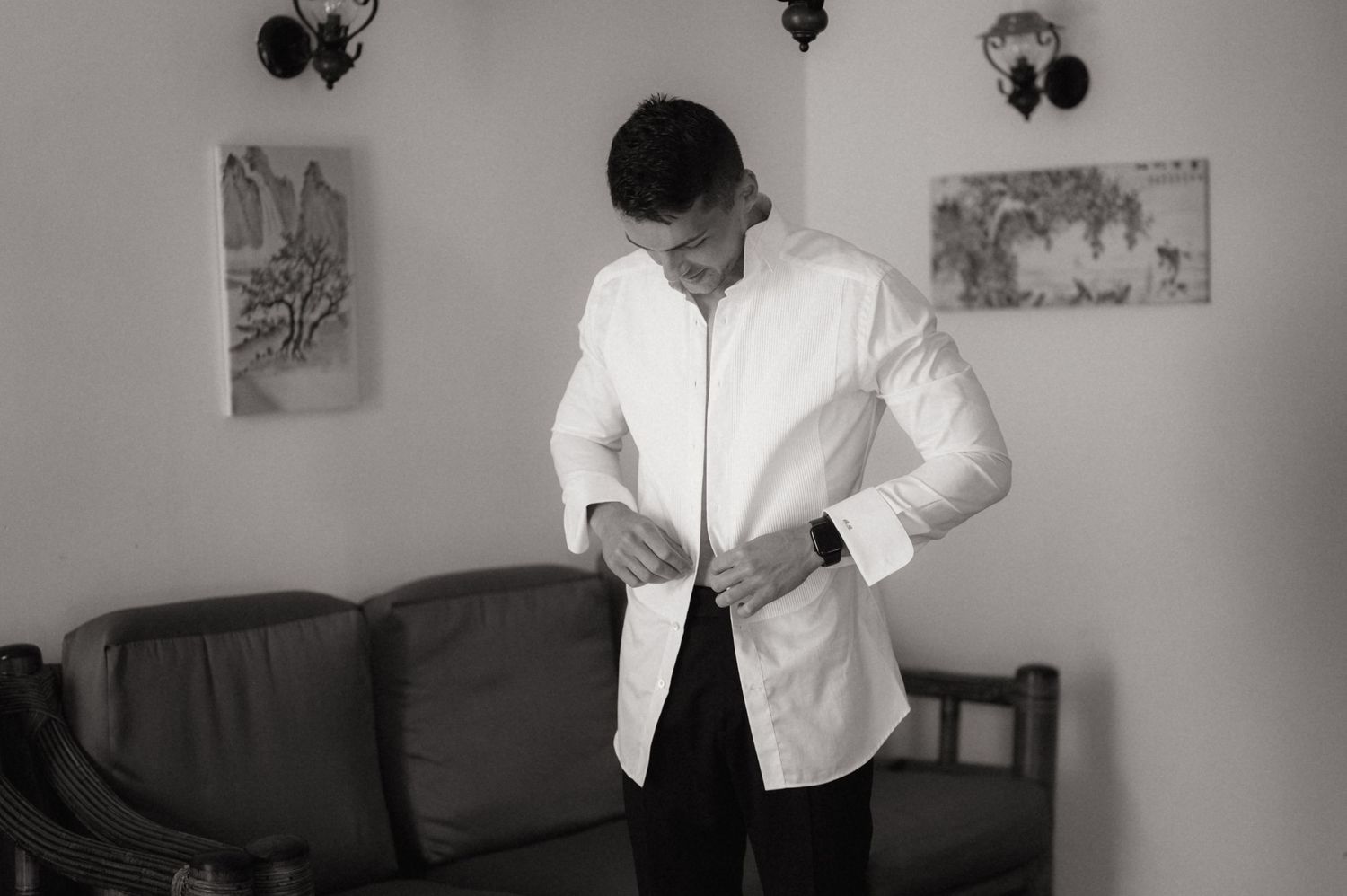 Black and white photo of a man in a white dress shirt buttoning up while standing in a living room.