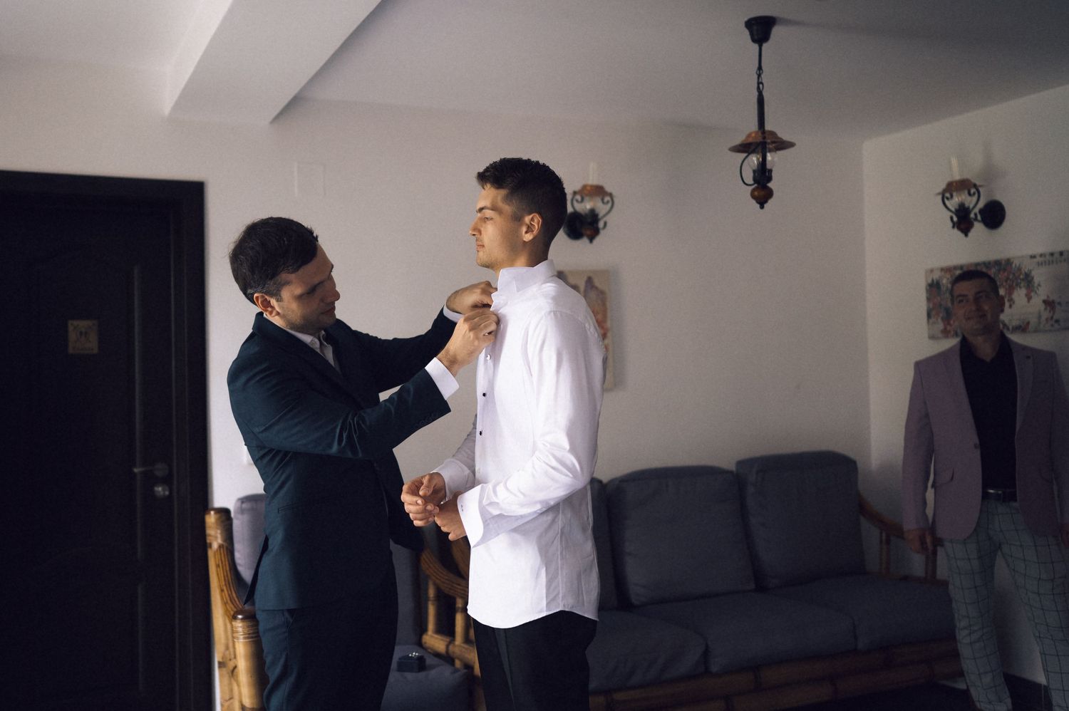 Man in suit adjusting tie of another man in white shirt, with a third man standing in background near sofa.