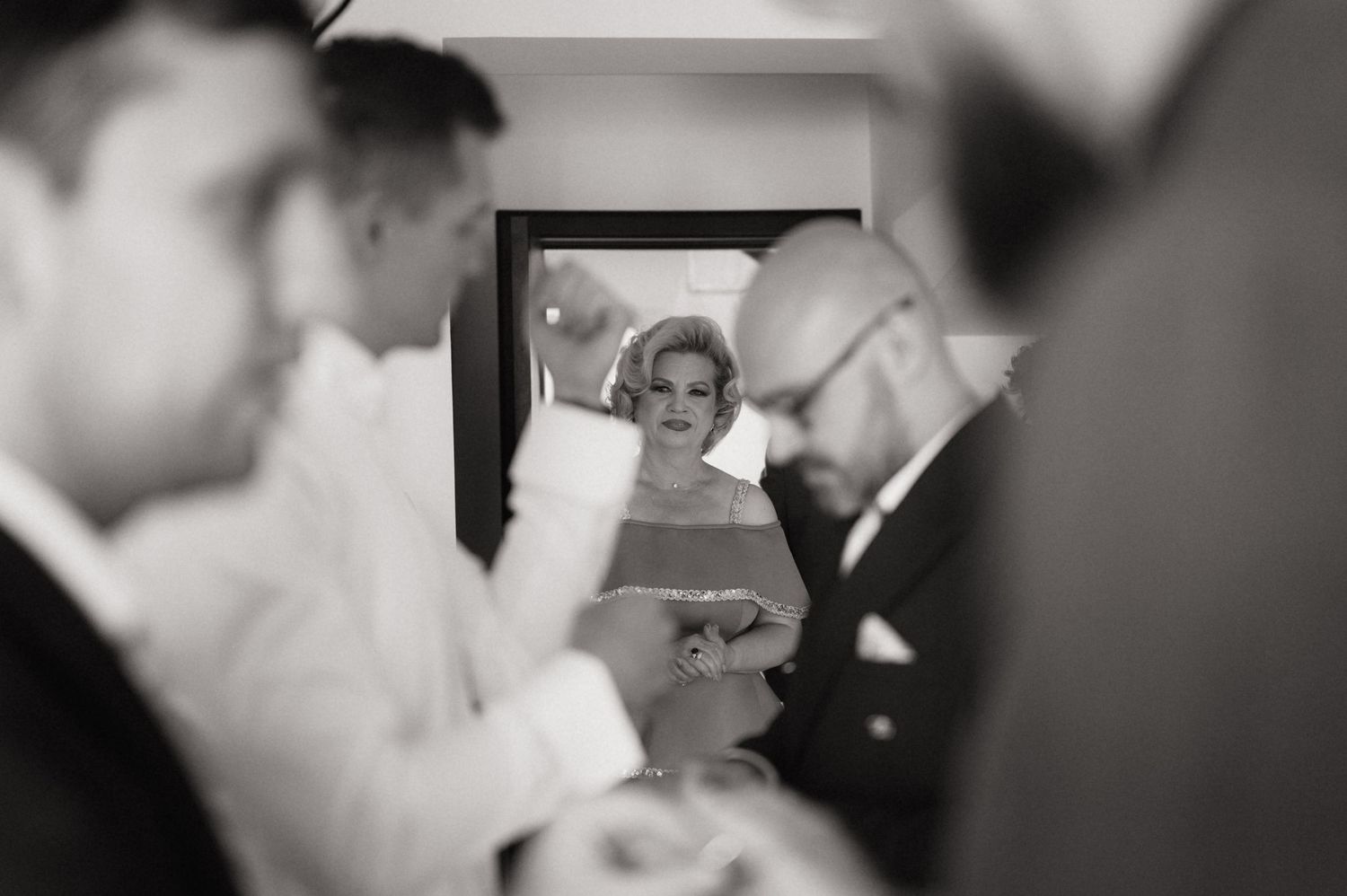Black and white wedding photo of a smiling woman in elegant attire, framed between blurred guests in formal wear.