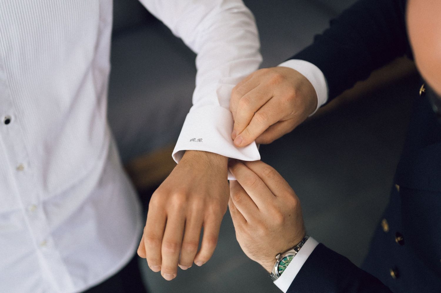 A person fastening cufflinks on a white dress shirt sleeve, assisted by another person in a suit.