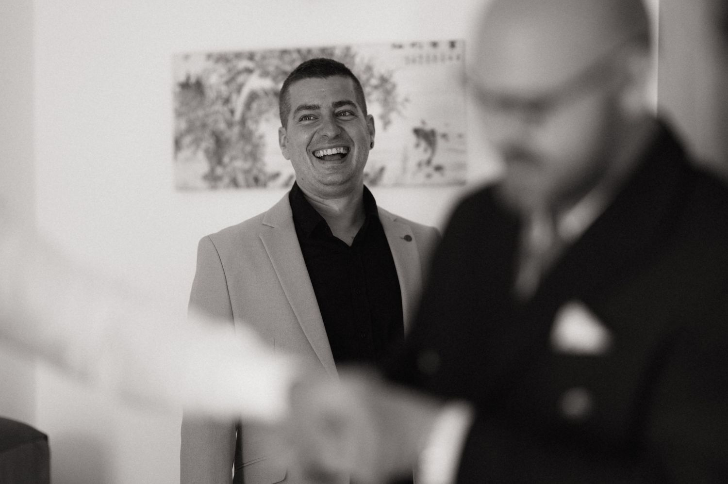 Black and white photo of a laughing man in a light blazer at a formal event with another man in foreground.