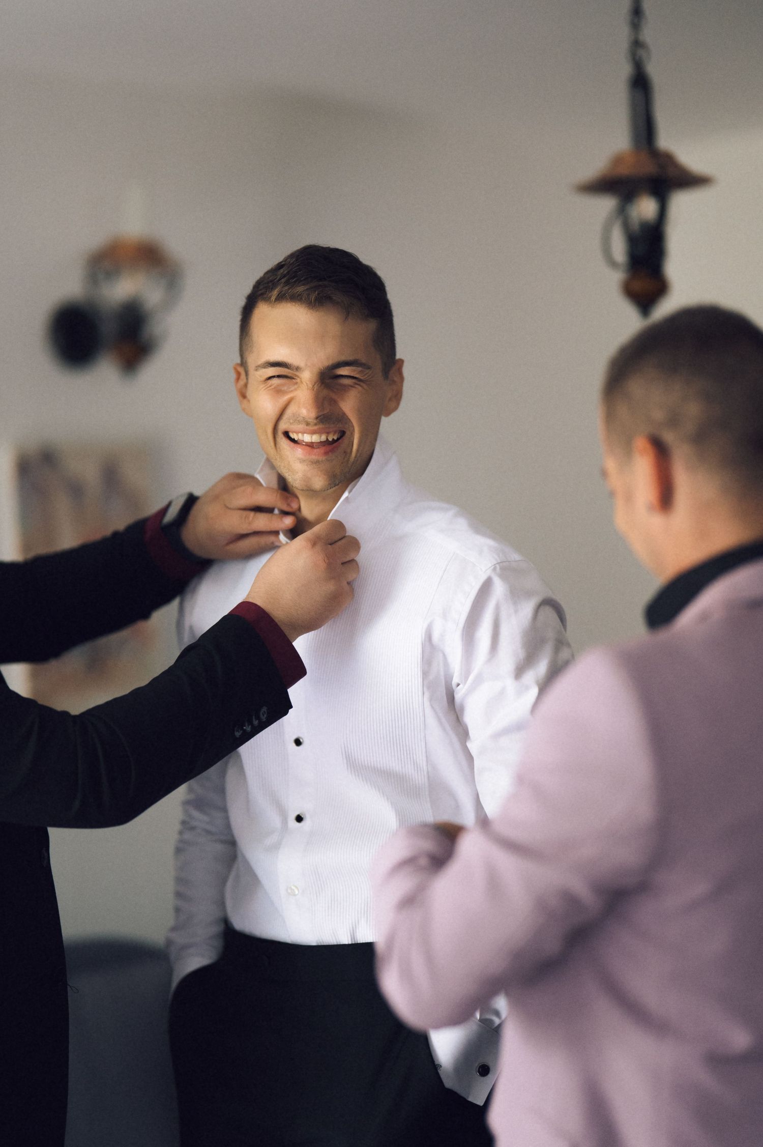 Smiling groom in white dress shirt gets help from groomsmen while preparing for his wedding day.