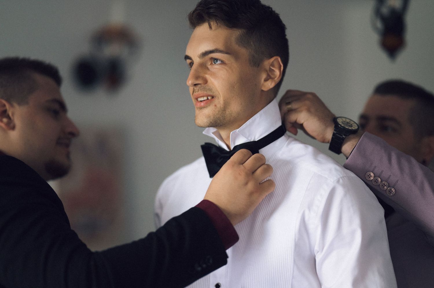 Groomsmen helping groom adjust black bow tie on white dress shirt before wedding ceremony.