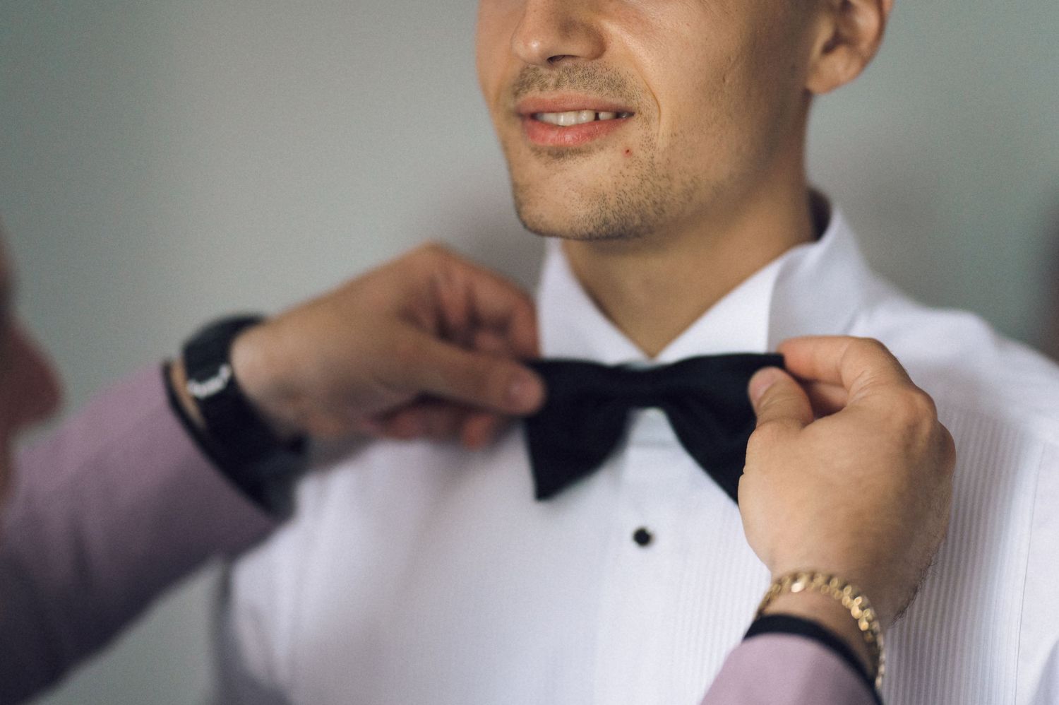 A groom in a white tuxedo shirt has his black bow tie adjusted by another person wearing a gold bracelet.