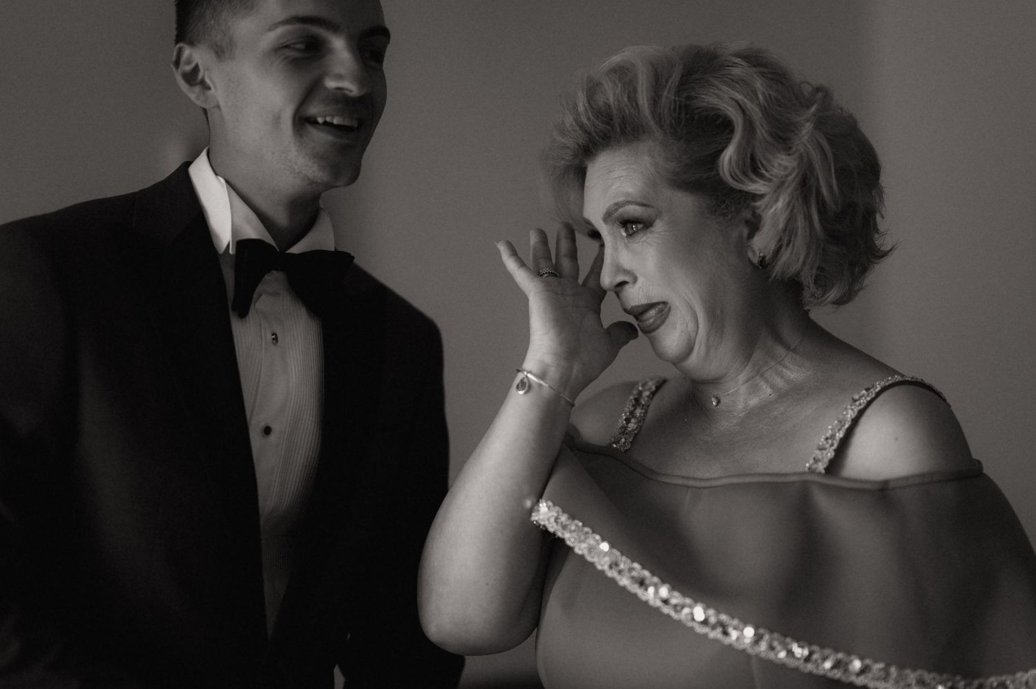 Black and white photo of a young man in tuxedo and elegant woman in gown whispering at formal event.