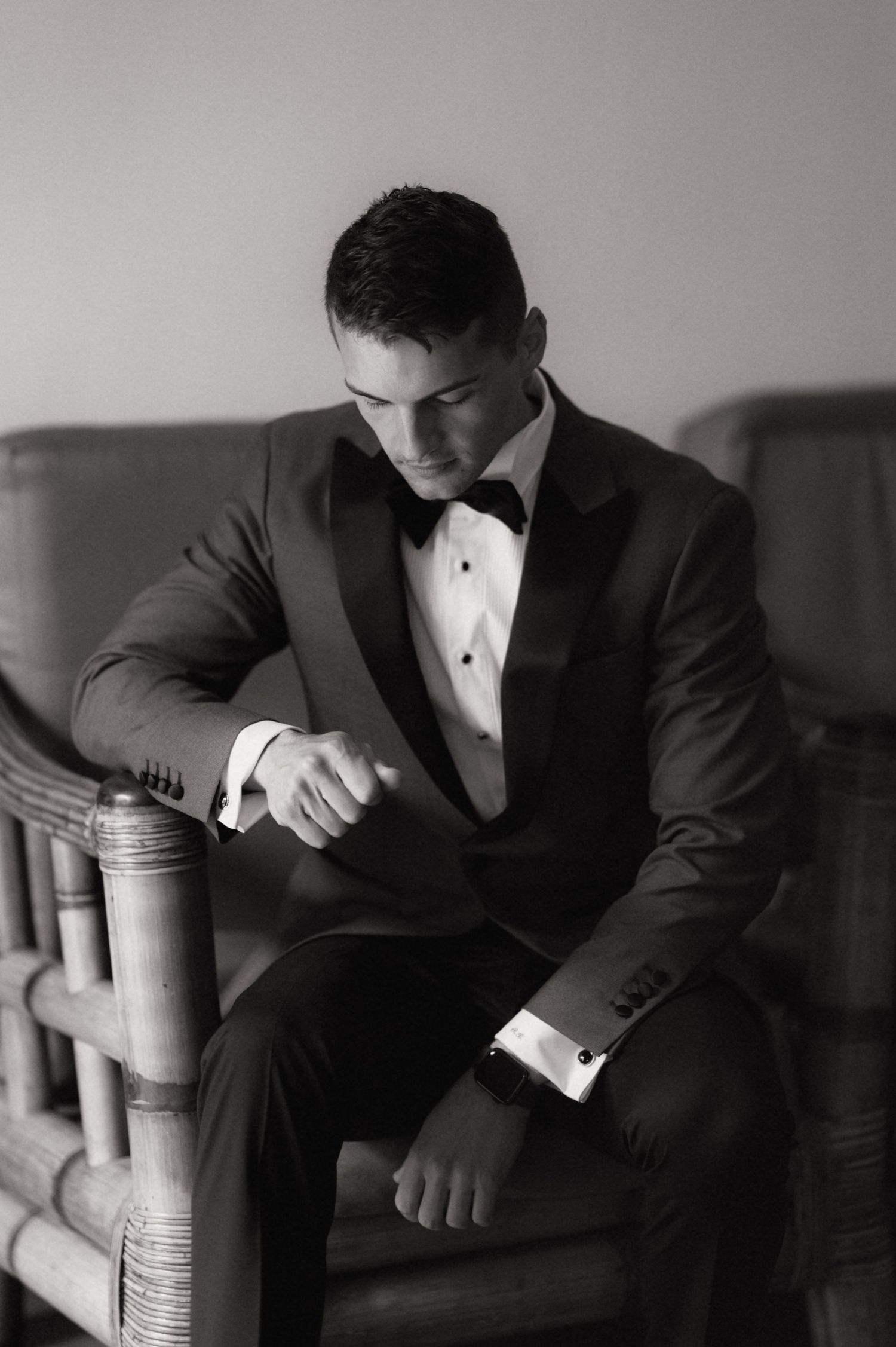 Black and white photo of a groom in a tuxedo and bow tie, sitting in a chair while checking his watch.