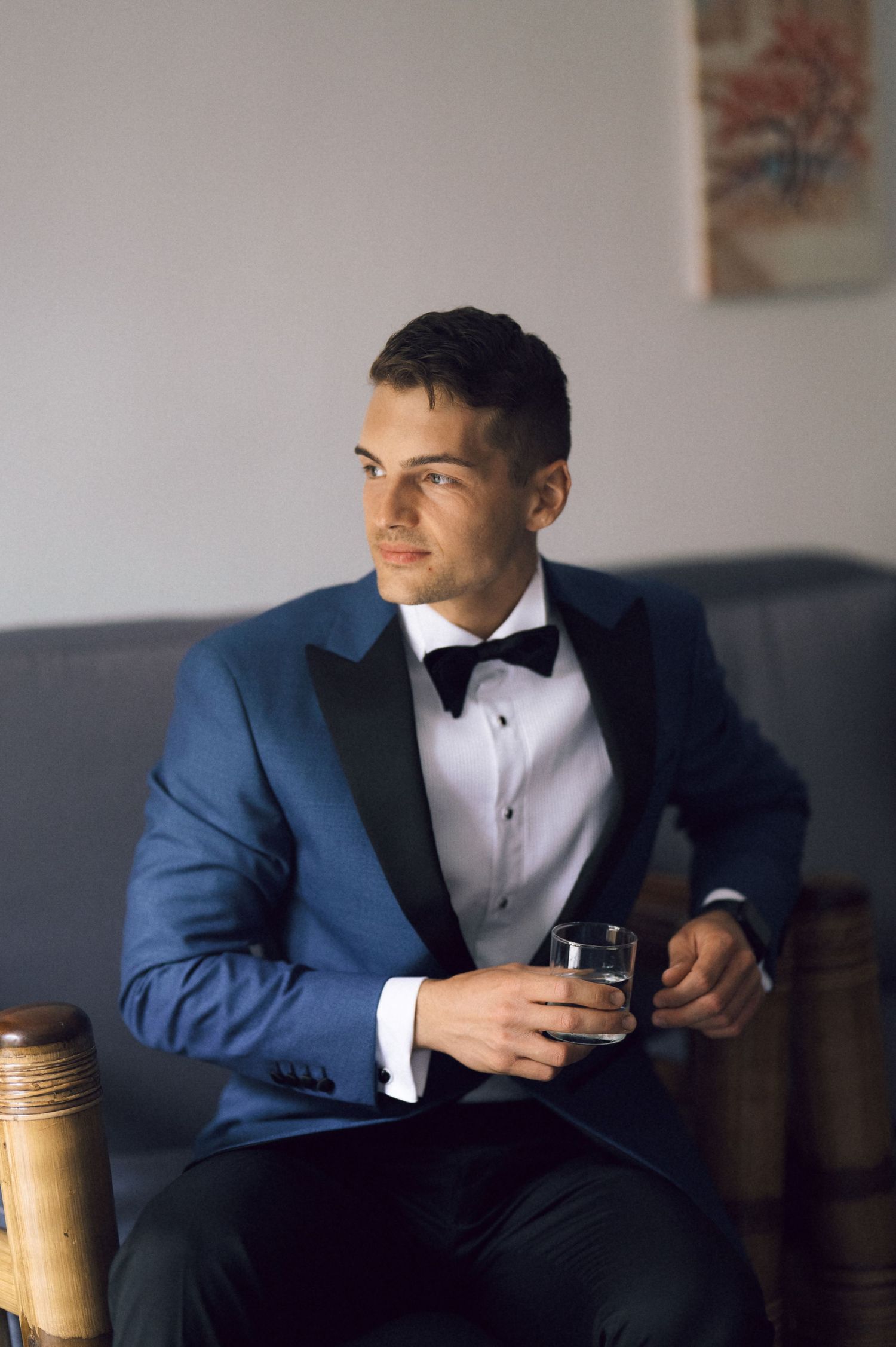 Elegant man in blue tuxedo with black lapels and bow tie, holding whiskey glass while seated.