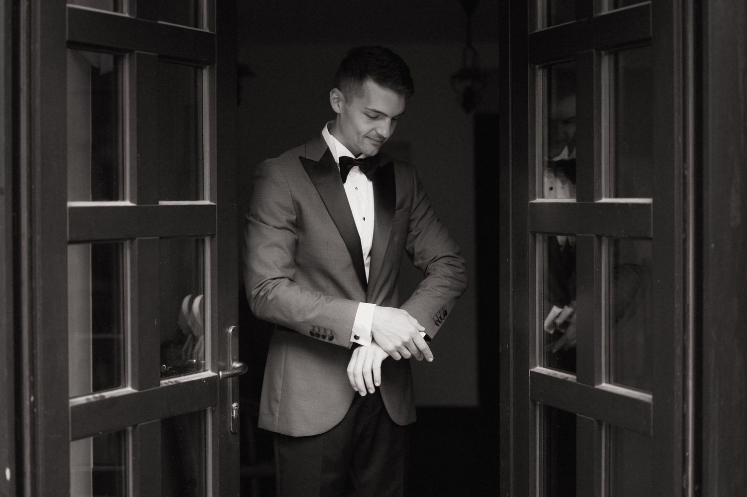 Black and white photo of a groom in a tuxedo adjusting his cufflinks in a doorway.
