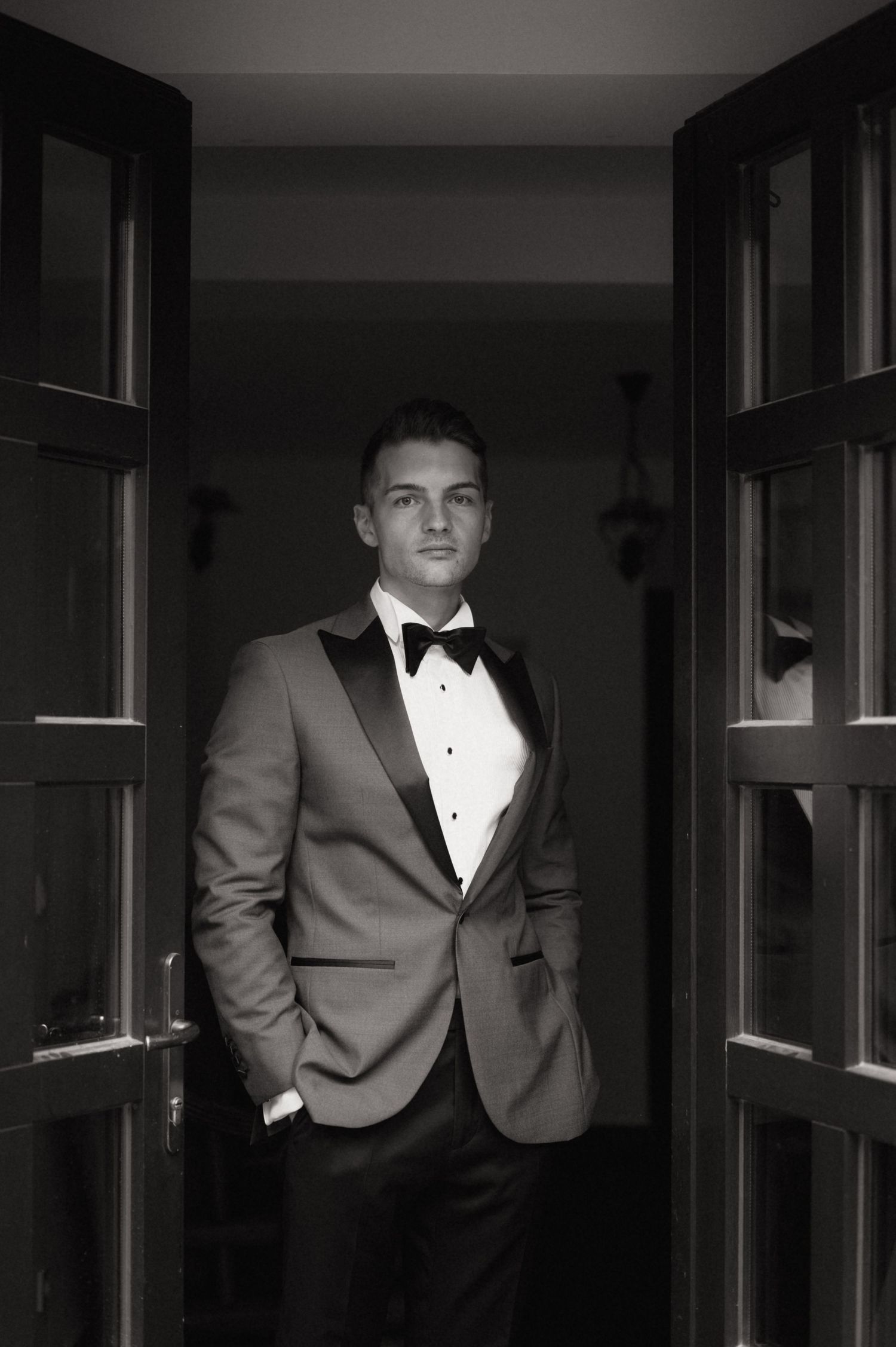 Black and white photo of a young man in a tuxedo with bow tie standing between double doors.