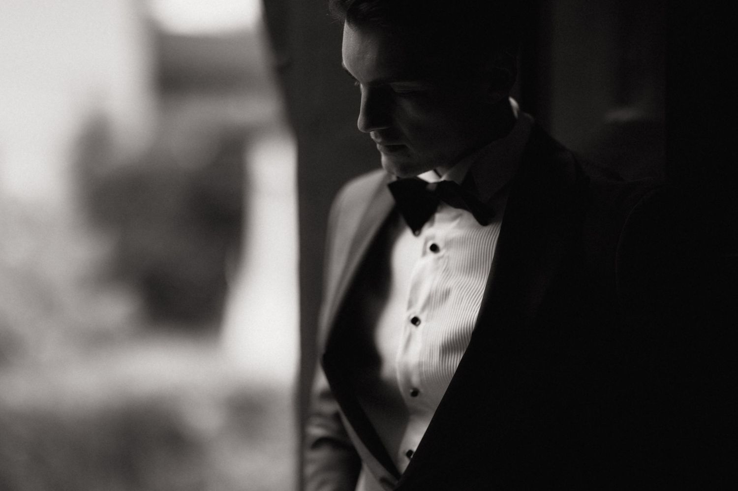 Black and white portrait of a man in a tuxedo with bow tie looking down in dramatic lighting.