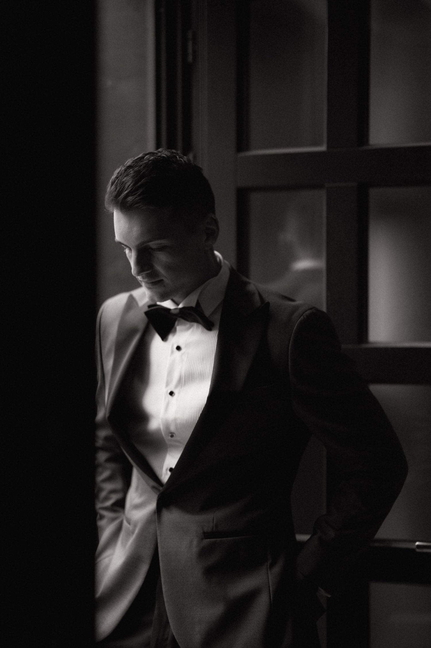 Black and white photo of an elegant man in a tuxedo with bow tie, standing by a window in dramatic lighting.