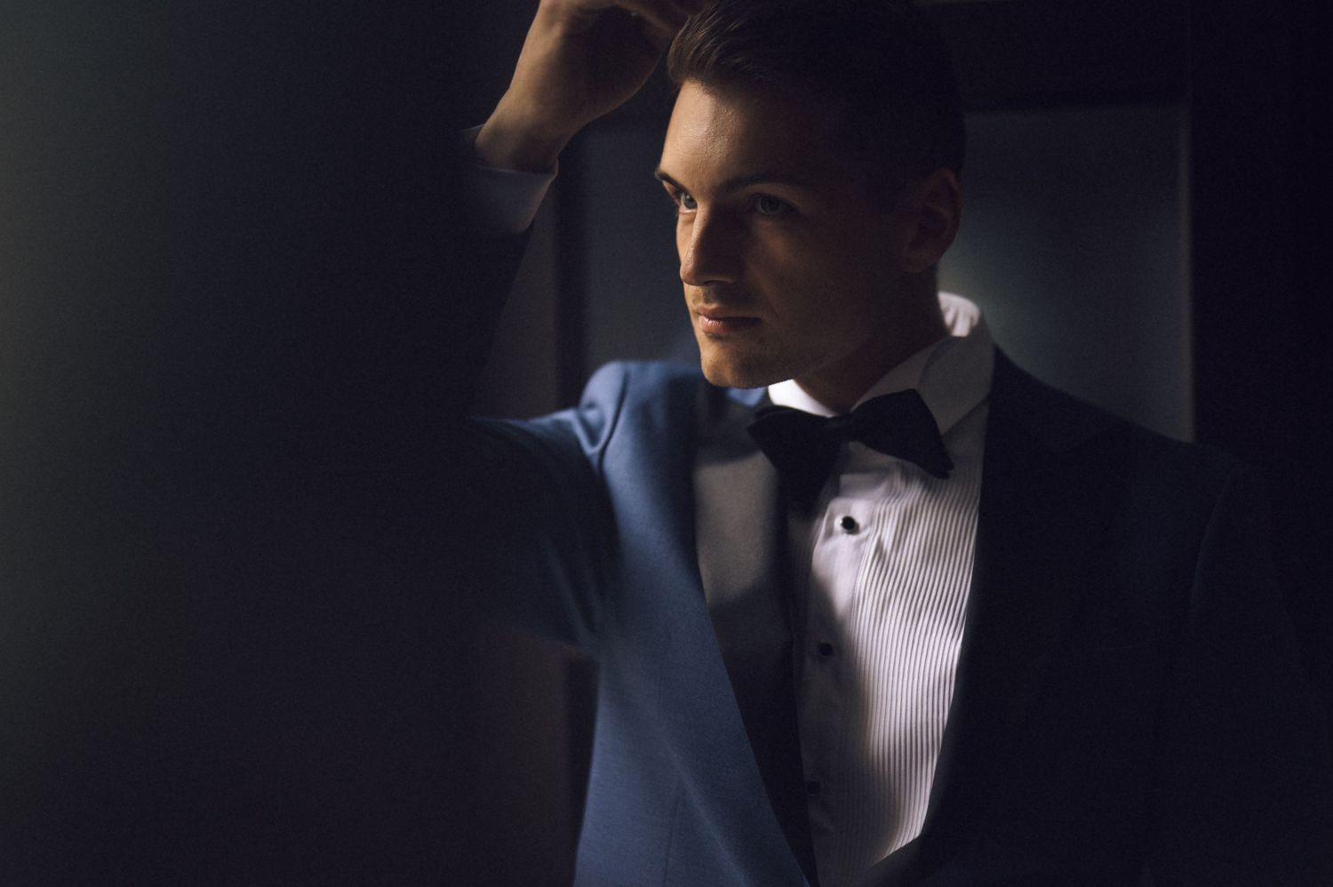 A sophisticated man in a black tuxedo with bow tie poses dramatically in moody lighting.