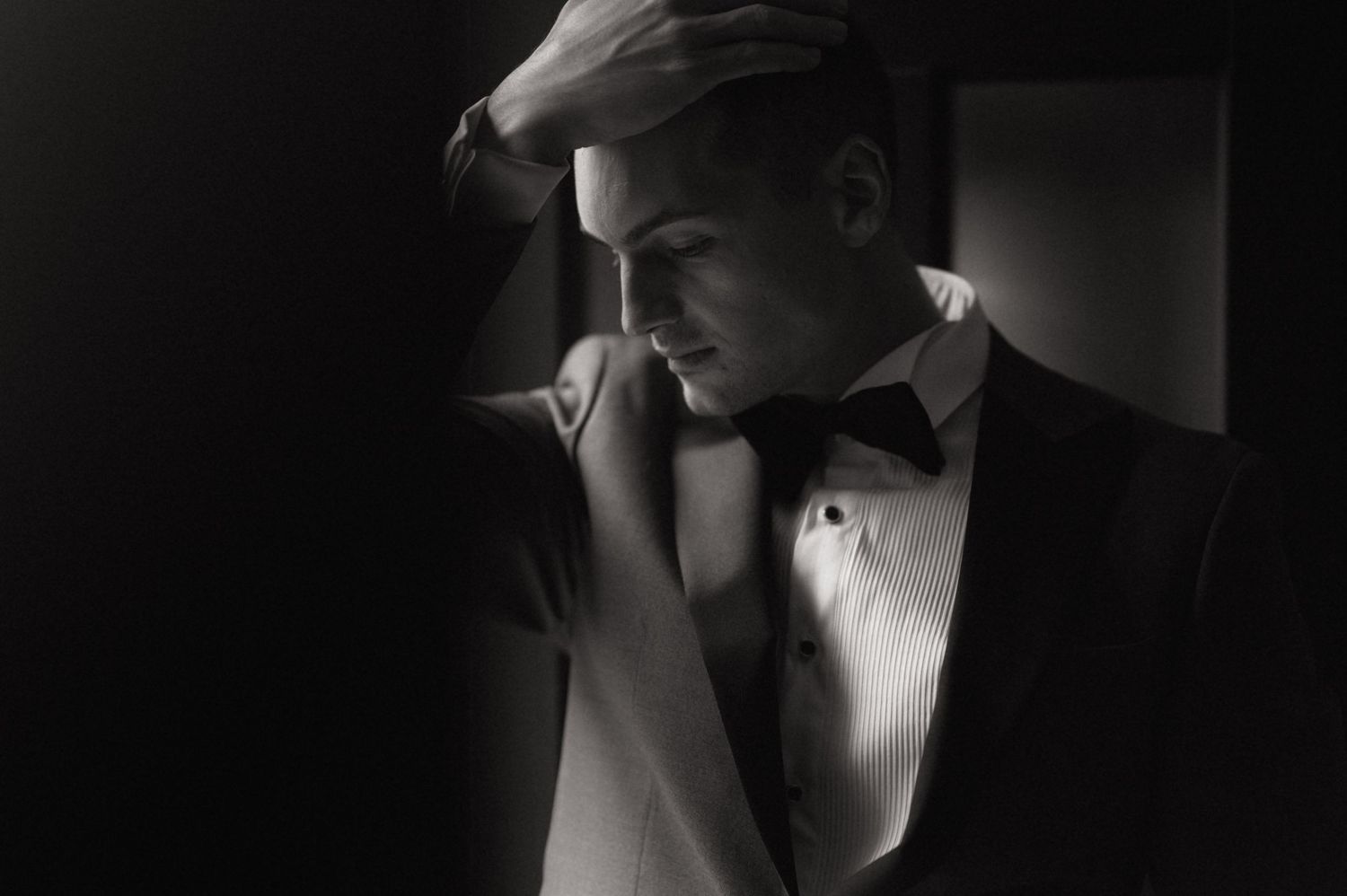 Black and white portrait of a man in a tuxedo with bow tie, hand raised to forehead in dramatic lighting.
