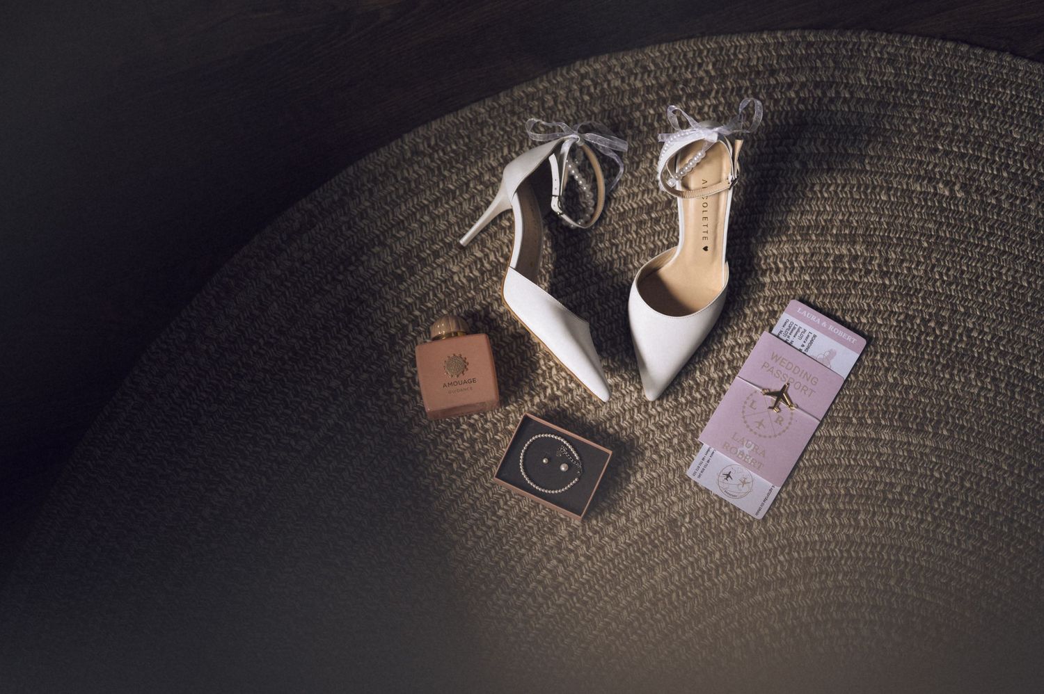 White bridal heels with lace-up ties displayed on woven surface beside passport, perfume, and pink ticket.