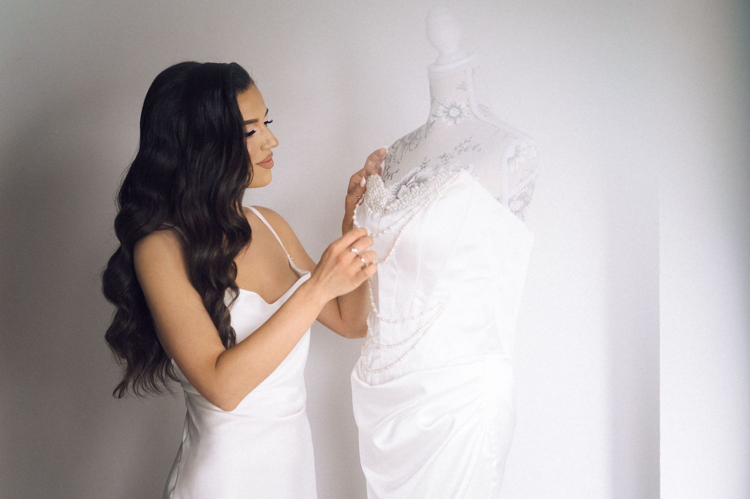 Bride in white gown examining elegant lace wedding dress on hanger against light gray background.