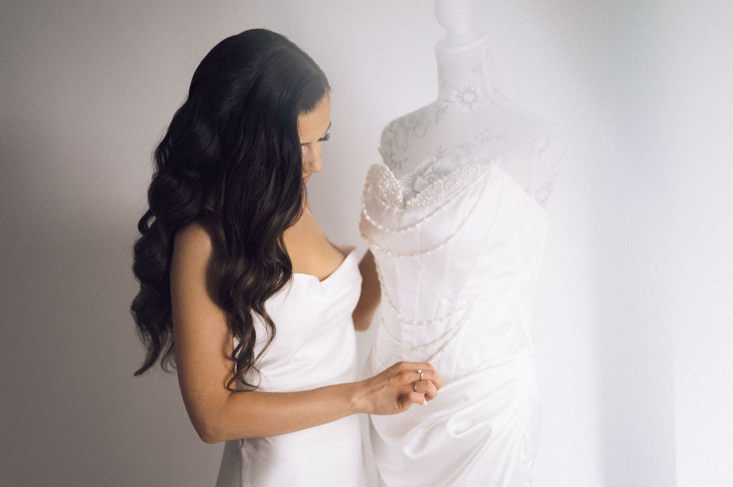 Bride with long dark hair holding a white lace wedding dress against a soft white background.