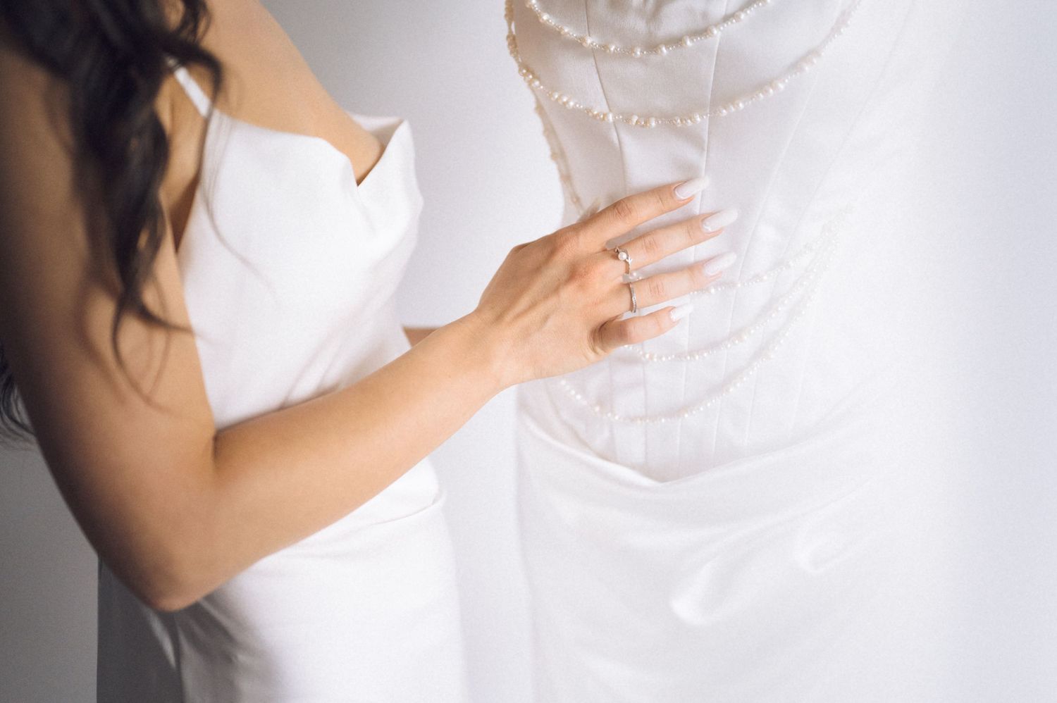 Bride in white dress touching pearl-adorned wedding gown, wearing engagement ring, elegant bridal preparation moment.