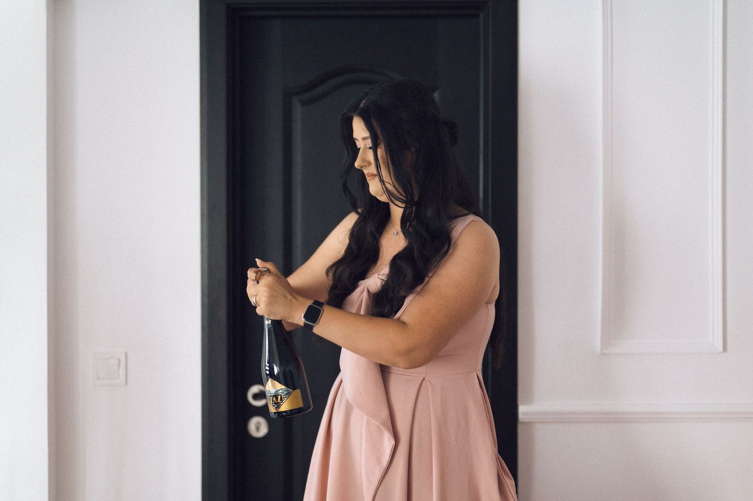 Woman in pink dress holding jewelry near a black door in an elegant white interior setting.