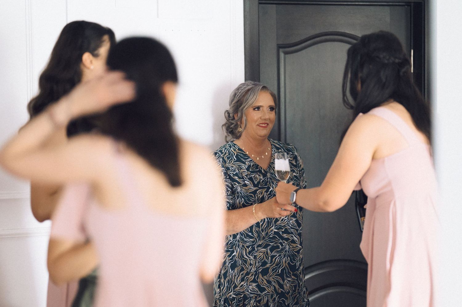Women in pink bridesmaid dresses chat with an older woman holding a wine glass near a dark door.