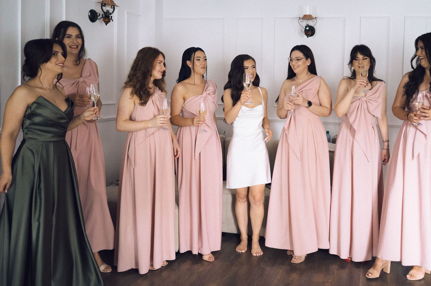 Bridesmaids in pink dresses holding champagne glasses stand with bride in white dress in elegant venue.