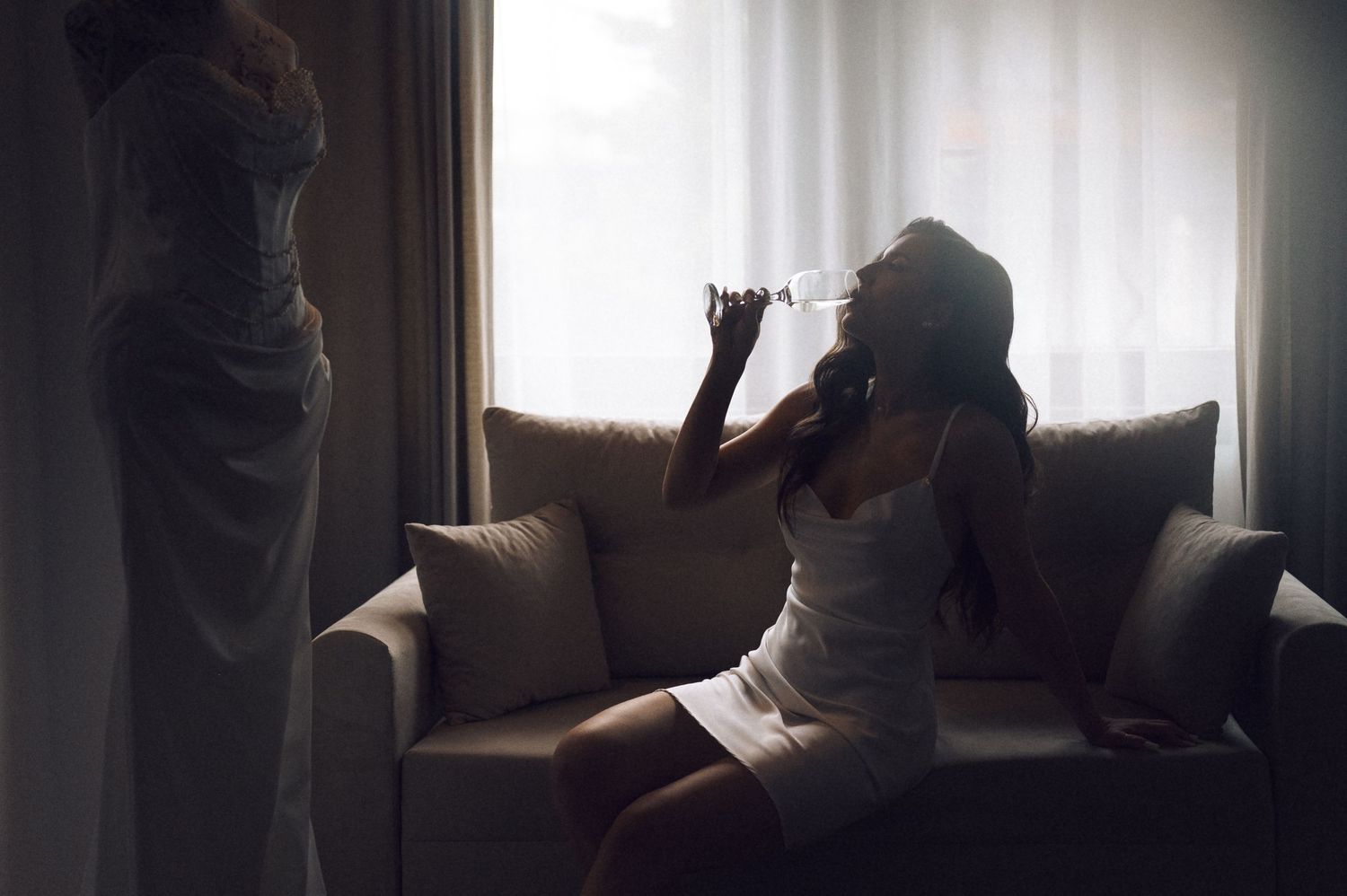 Woman in white dress sitting on sofa sipping champagne near a wedding gown by a bright window.