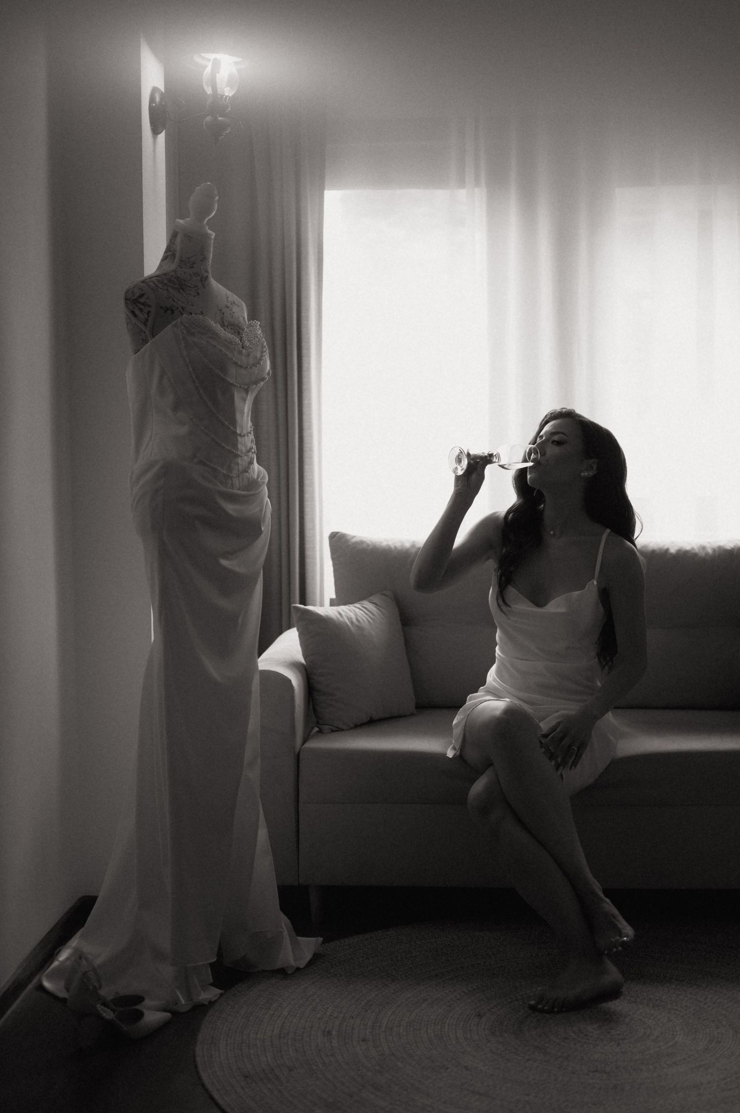 Bride in white slip dress sips champagne beside her wedding gown hanging by a sunlit window, black and white photo.