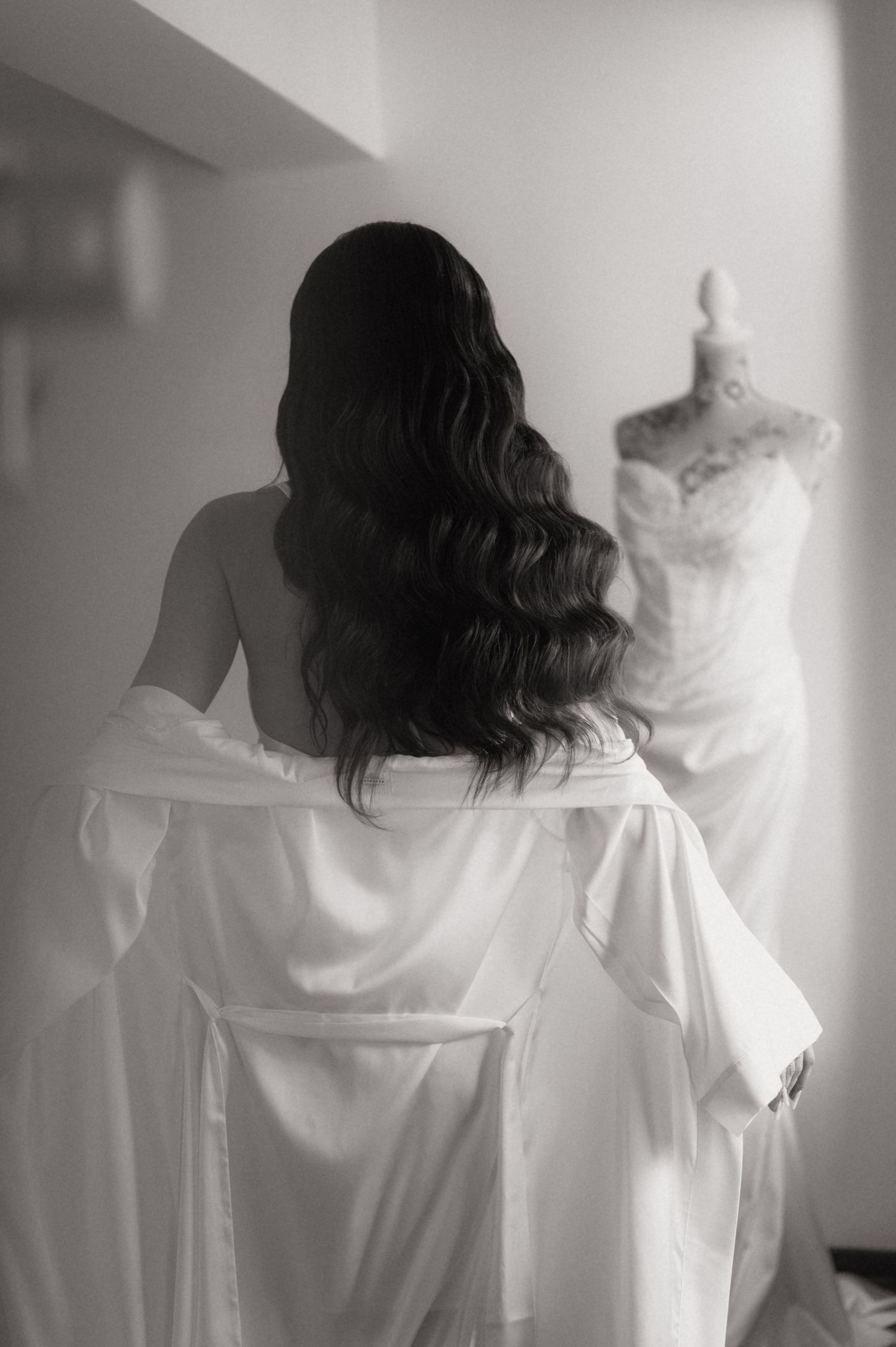 Bride in white robe with long wavy hair gazes at her wedding gown on a mannequin in a black and white photo.