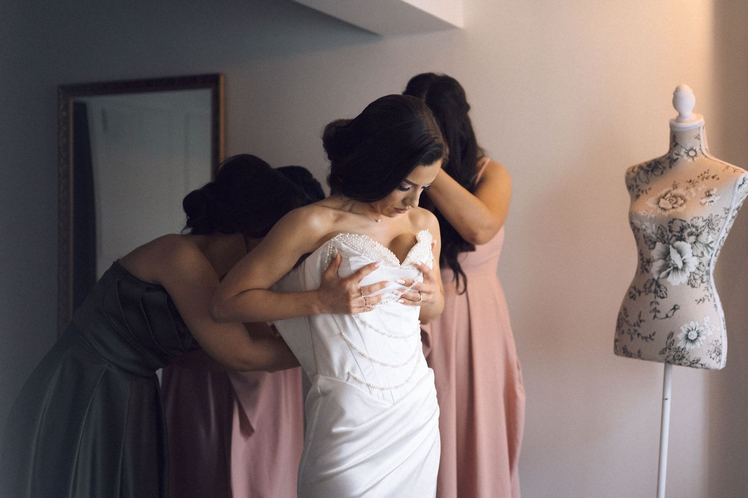 Bridesmaids helping bride into white wedding dress near a dressmaker mannequin in a softly lit bridal suite.