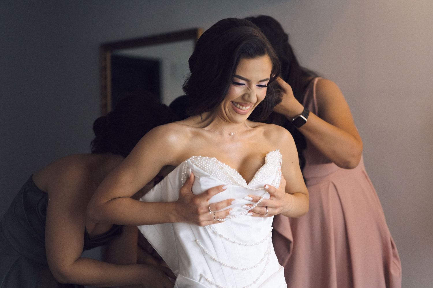 Bride smiling joyfully as bridesmaids help her into a white strapless wedding gown before the ceremony.