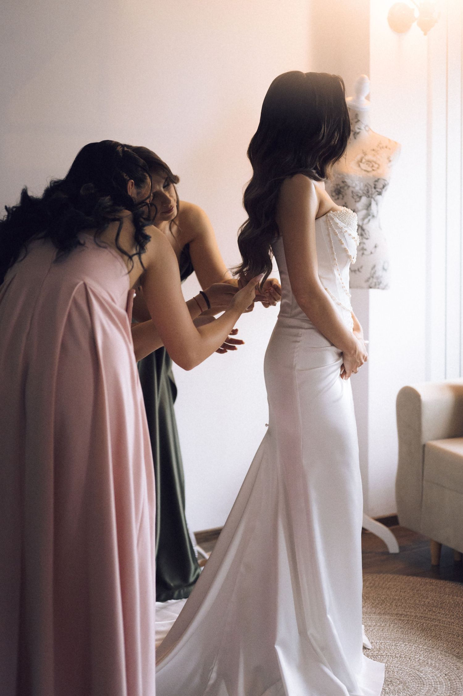 Bridesmaids in pink and green gowns helping bride into fitted white wedding dress in elegant bridal suite.