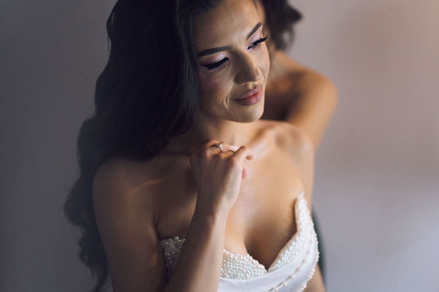 Elegant bride in white lace gown smiling softly, touching her necklace with flowing dark hair.