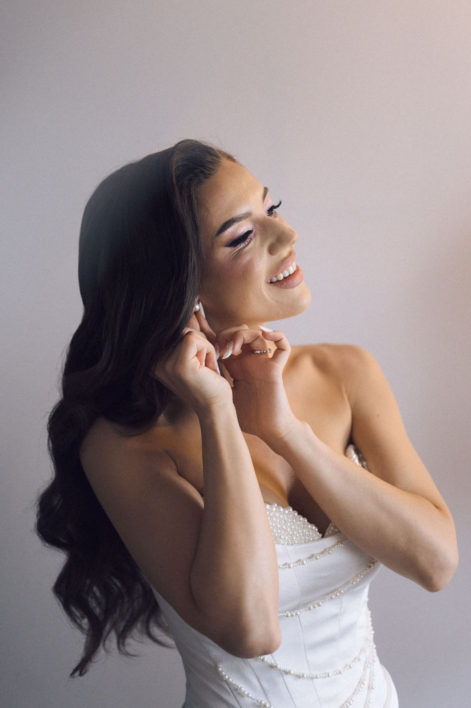 Smiling bride with dark wavy hair adjusting earring in white strapless beaded wedding gown.