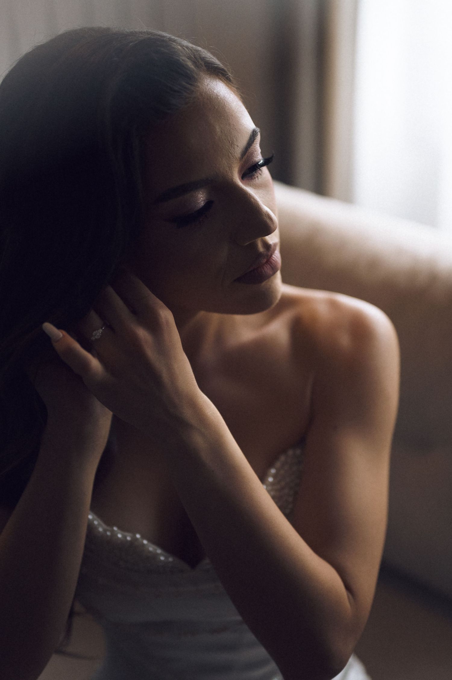 Elegant bride with dark hair adjusting her hair, wearing a pearl-embellished strapless wedding gown in soft light.