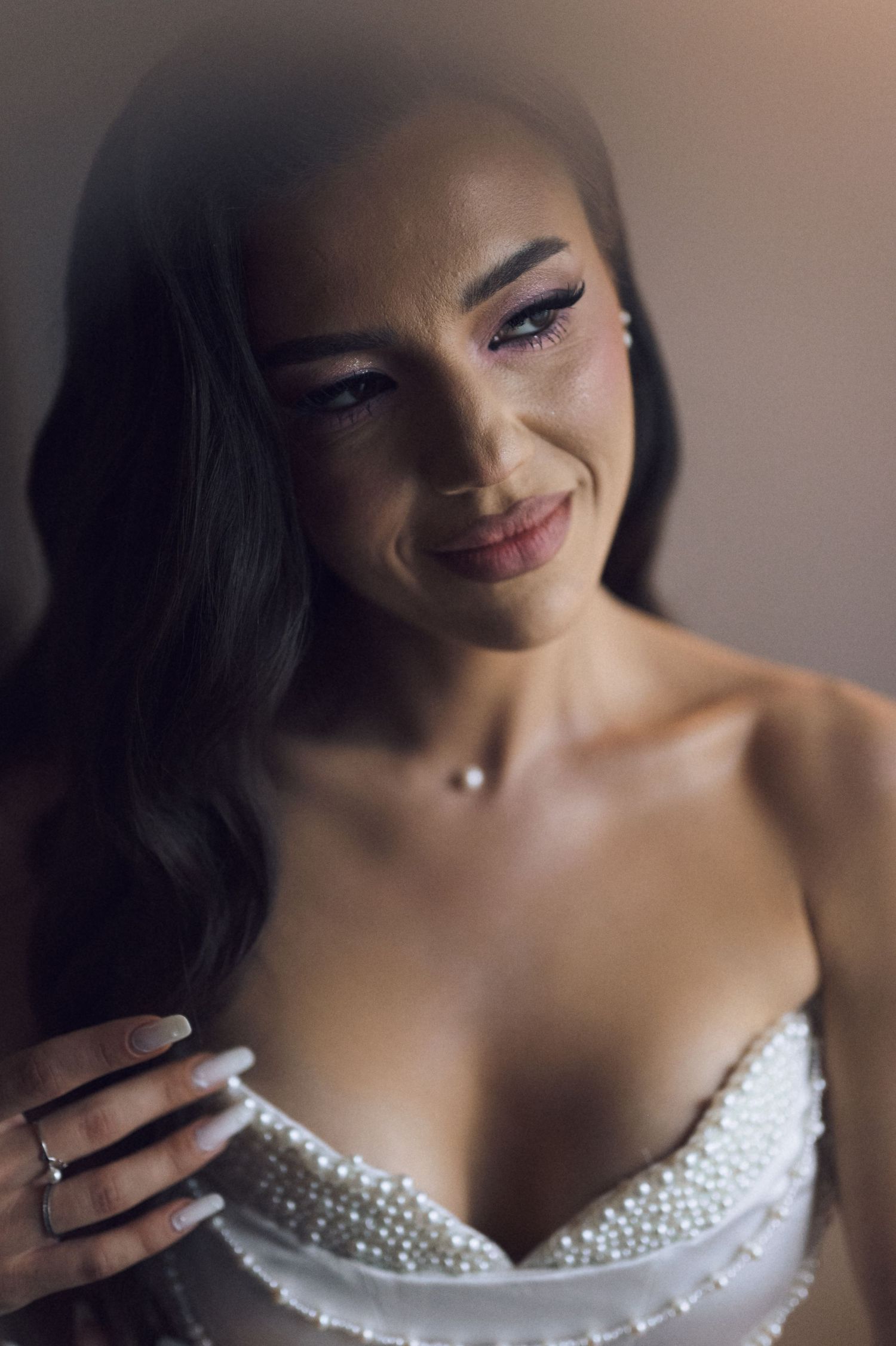 A bride in a pearl-embellished wedding gown smiles softly, wearing elegant makeup and a delicate diamond necklace.