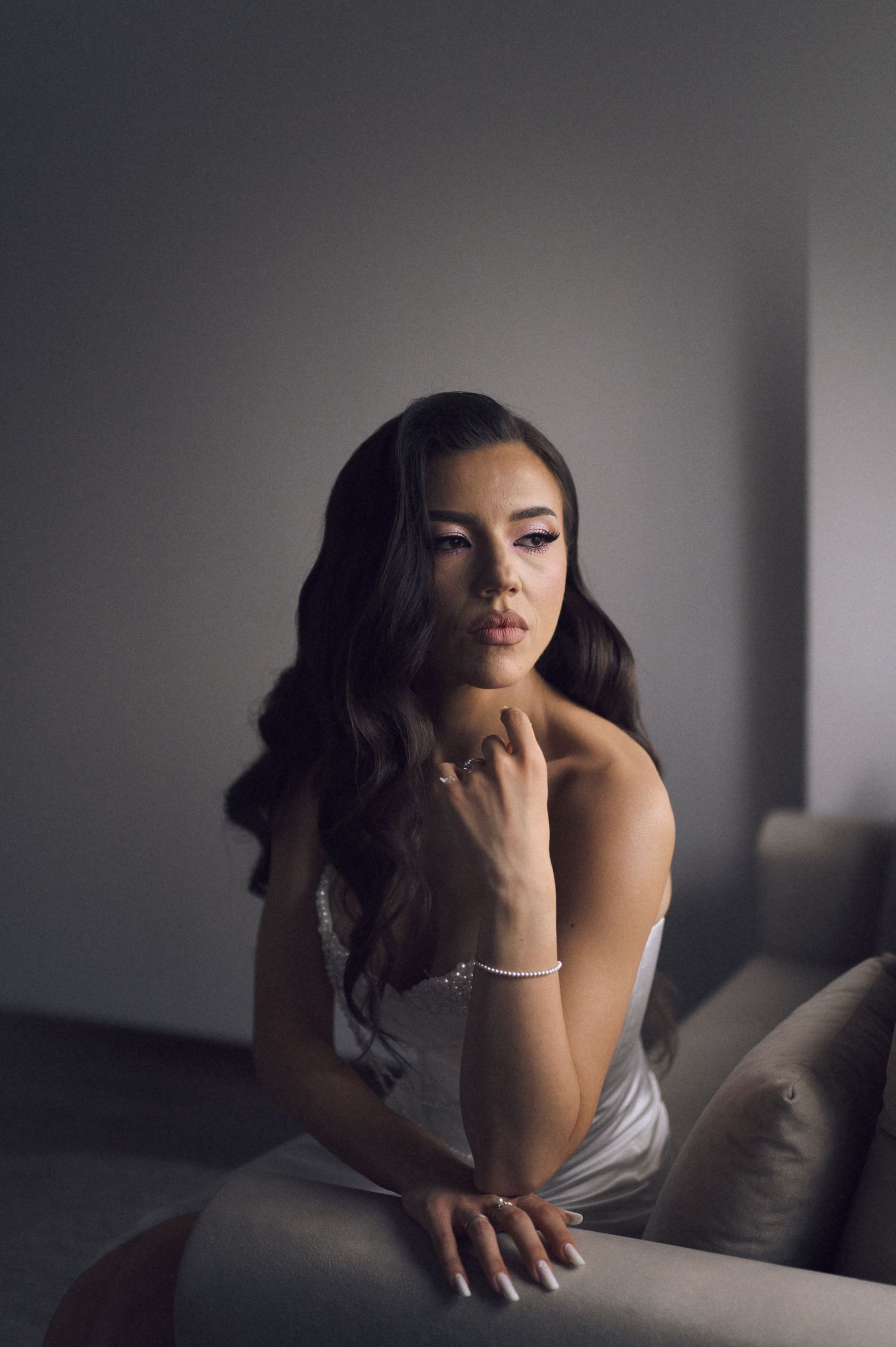 Young woman in elegant white dress with wavy dark hair and bracelet poses gracefully on a couch in moody lighting.