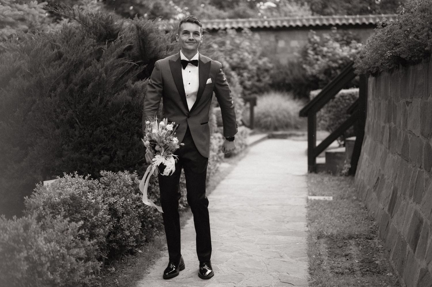 Groom in black tuxedo and bow tie walks garden path carrying bridal bouquet in elegant black and white wedding photo.