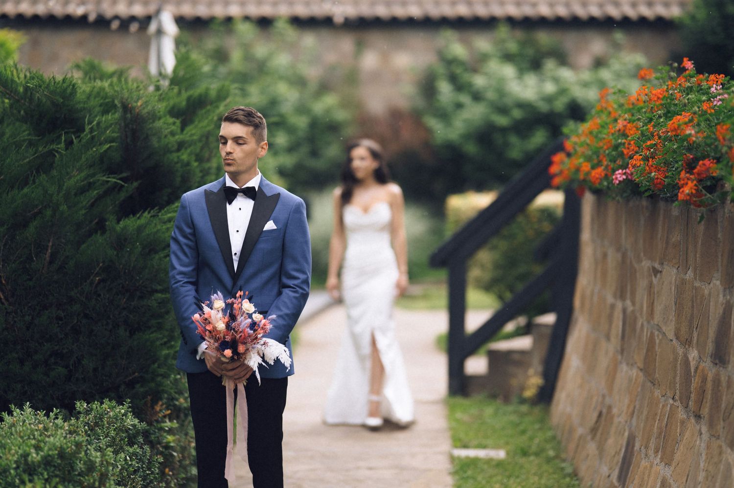 Groom in navy tuxedo holds bouquet as bride in white gown approaches on garden pathway with orange flowers.
